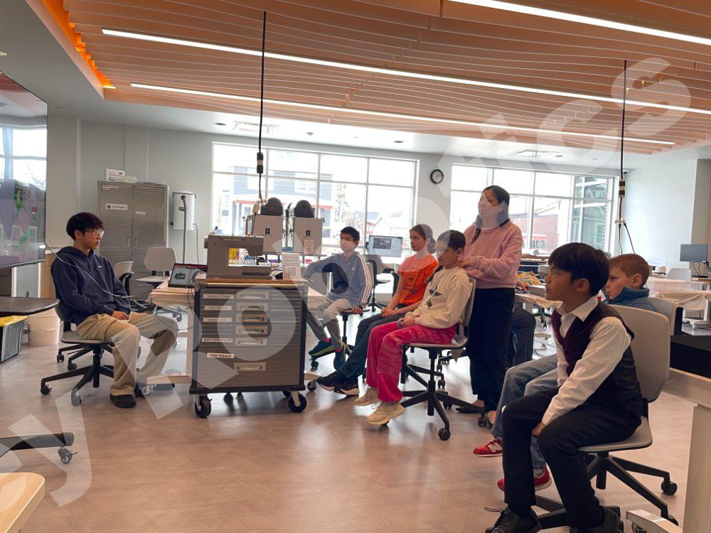 A student coach presenting a video on a large screen to students during a 2025 Fall Program class in a modern, bright robotics classroom.