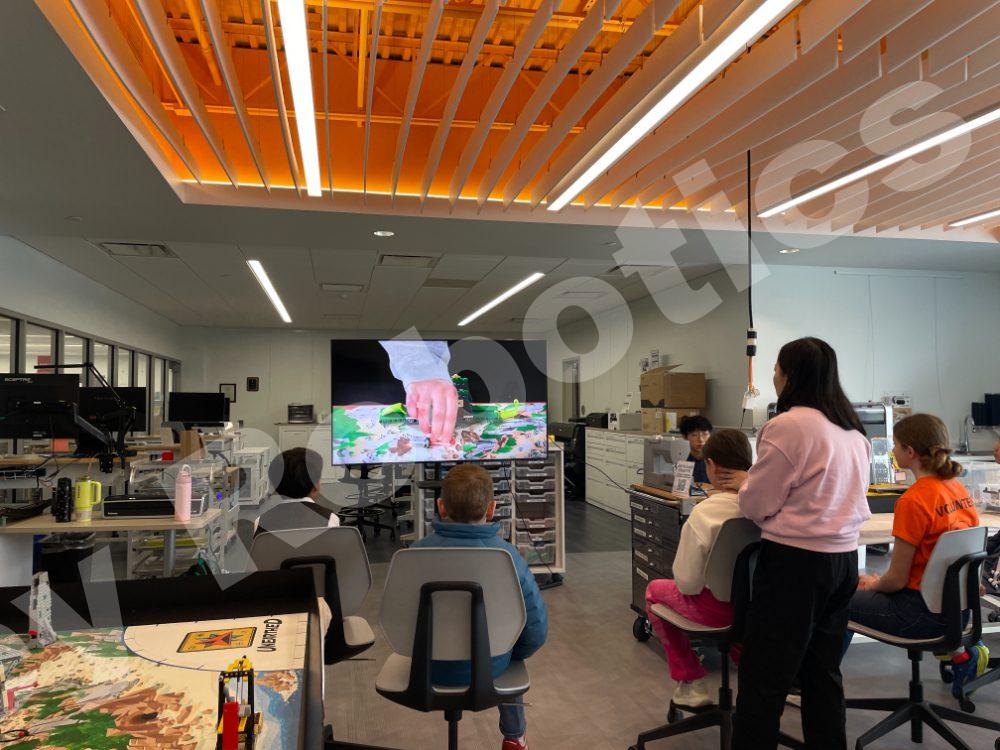A student coach presenting a video on a large screen to students during a 2025 Fall Program class in a modern, bright robotics classroom.