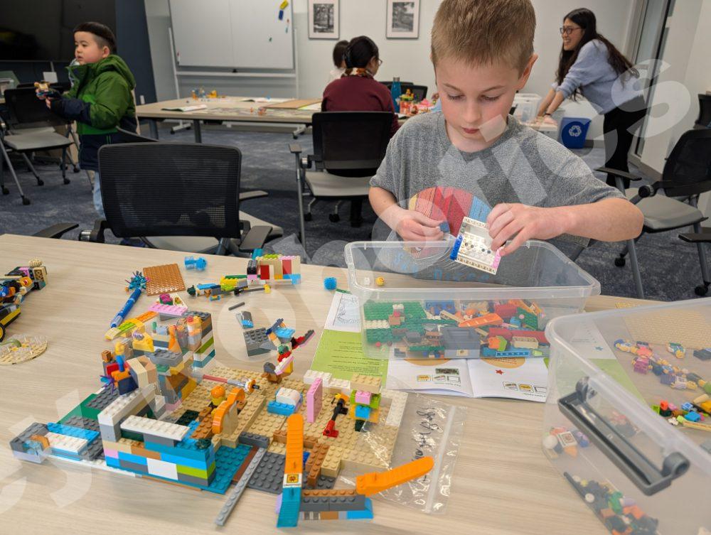 An elementary student carefully examines Lego pieces while a student coach works with other children in the background during a 2025 Fall Program class.